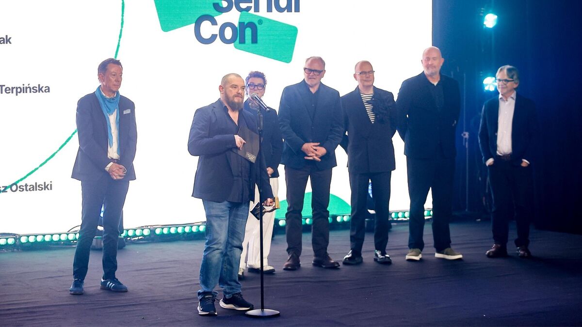 Grupa siedmiu osób stoi na scenie podczas Warsaw Serial Con, a mężczyzna z brodą przemawia do mikrofonu, trzymając w ręku kartkę lub kopertę. Pozostali uczestnicy ustawieni są w półokręgu za nim, w stonowanych strojach; po lewej stronie widać mężczyznę w marynarce z niebieską apaszką. W tle świeci duży ekran z logotypem wydarzenia oraz fragmentami nazwisk, a po prawej stronie scenę doświetlają reflektory.