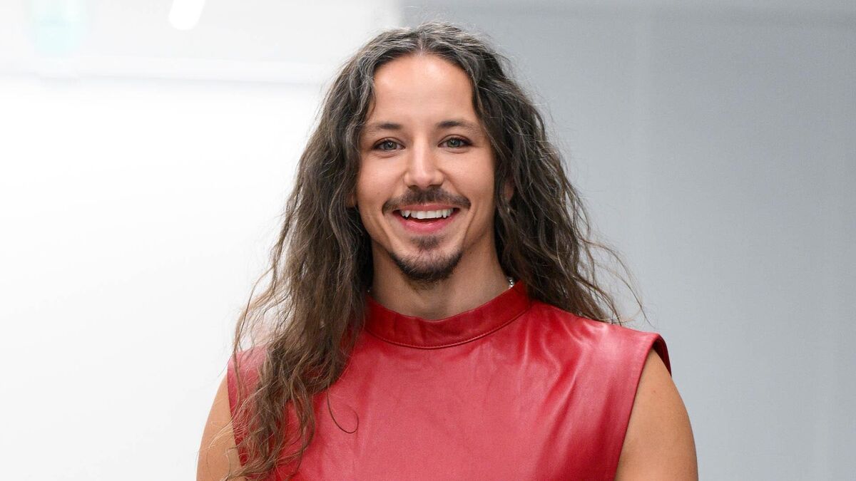 Michał Szpak pozuje do zdjęcia w czerwonej, skórzanej koszulce bez rękawów. Ma długie, falowane włosy oraz zarost z wąsami i kozią bródką. W tle znajduje się jasne, rozmyte wnętrze z białymi i szarymi ścianami.