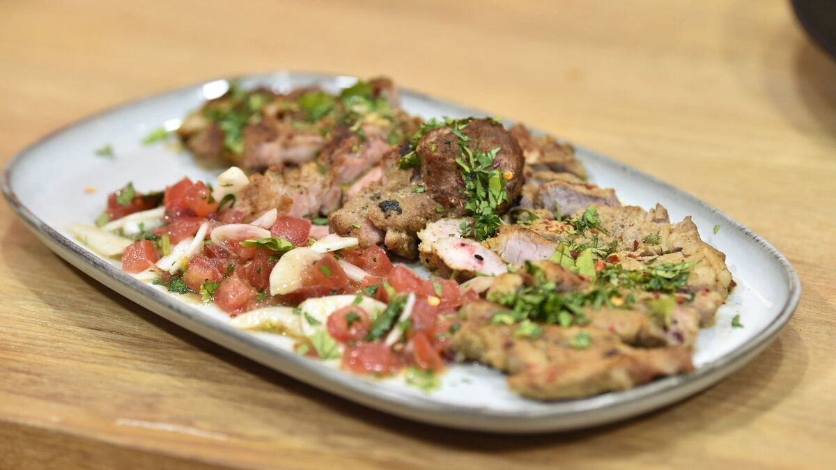 Karkówka Carne Asada jest podana na podłużnym talerzu, pokrojona na kawałki i posypana świeżymi ziołami. Obok mięsa znajduje się salsa z pomidorów i czosnku, wymieszana z zielonymi dodatkami. Całość leży na drewnianym blacie, a tło jest rozmyte.