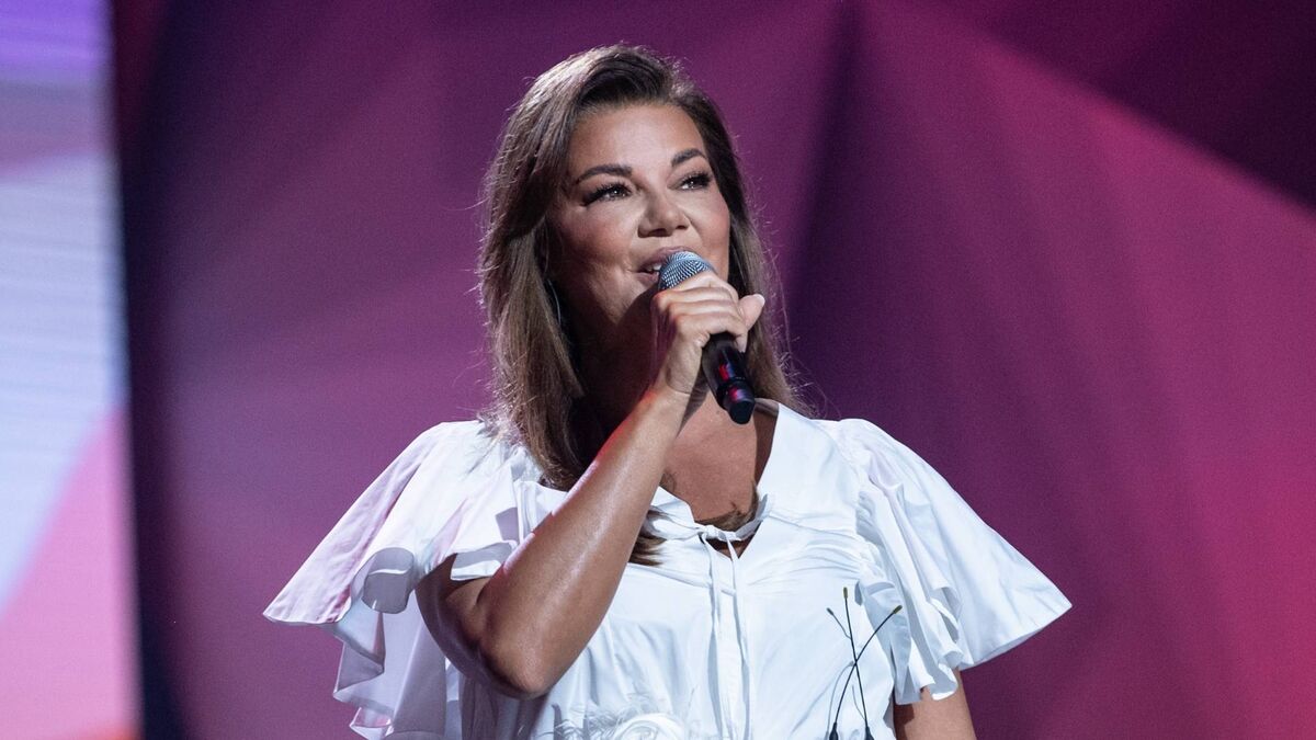 Edyta Górniak występuje na scenie, trzymając mikrofon blisko ust. Ma na sobie białą suknię z krótkimi, falbaniastymi rękawami, a tło oświetlone jest różowo-fioletowym światłem scenicznym. Włosy ma rozpuszczone do ramion, a fotografia została wykonana podczas koncertowego występu.